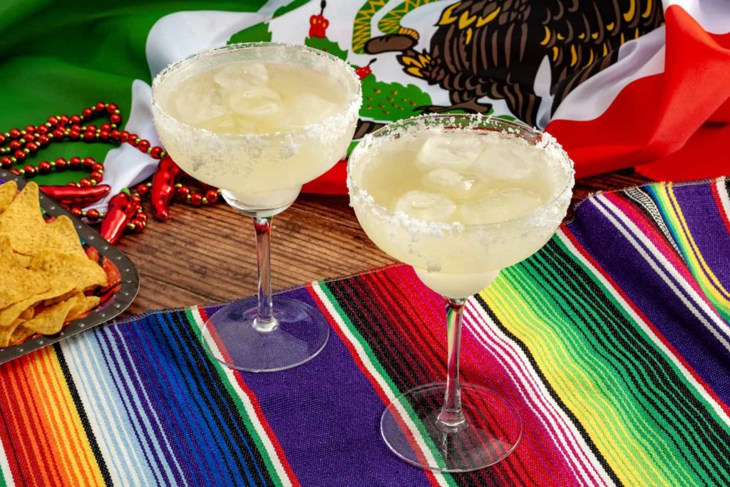 Two margaritas on a colorful placemat with a Mexican flag in the background.