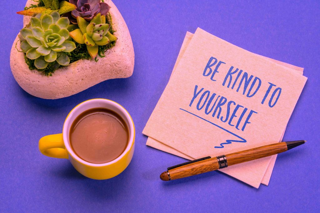 A coffee cup and a plant on a desk with a note reading, “be kind to yourself.”