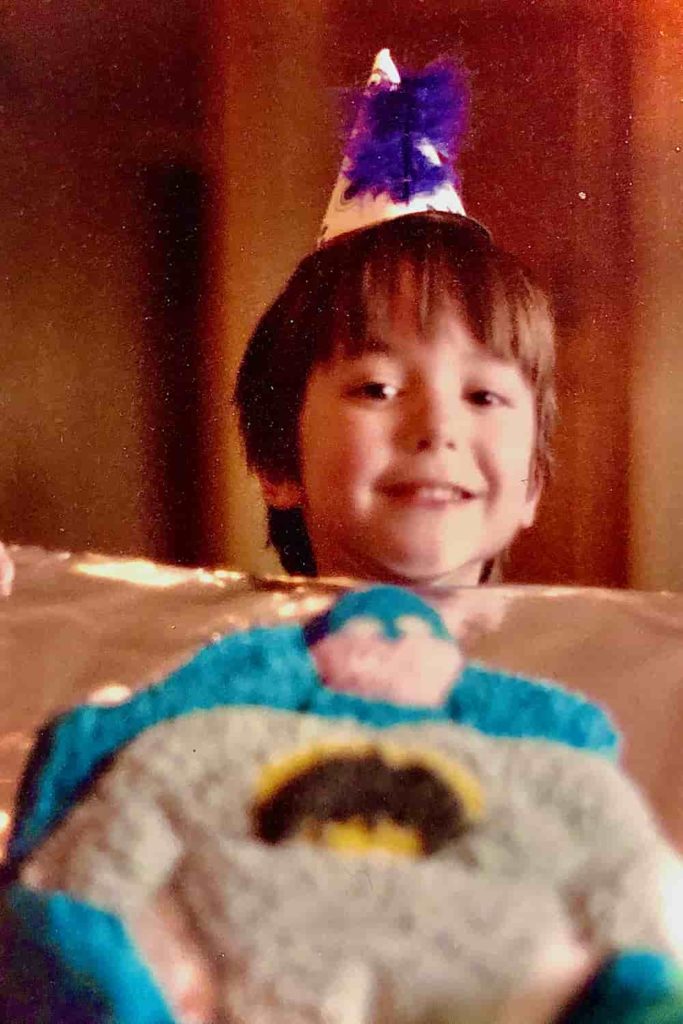 A little boy wearing a party hat posing behind a Batman birthday cake.