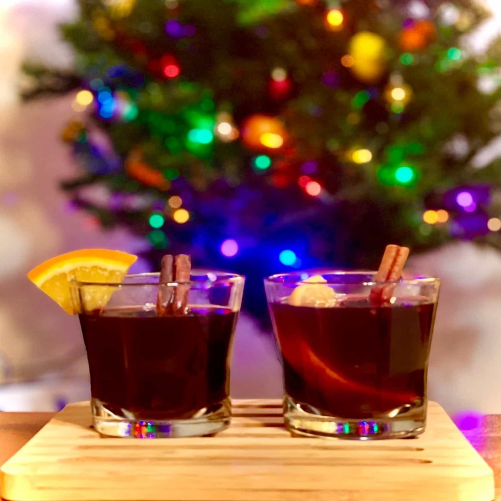 Two glasses of gluhwine with citrus garnish in front of an illuminated Christmas tree.