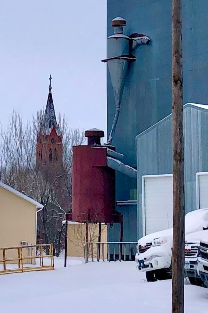 A photo of the back side of a farmer’s elevator with a church steeple in the background.