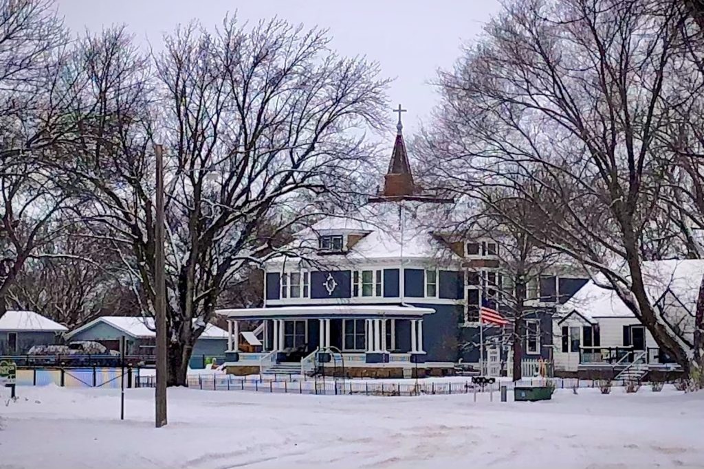 A photograph of a large house on a snowy day with a church steeple in the background.