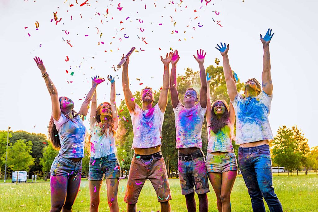 Six paint-covered people stretch their arms to the sky as confetti falls.