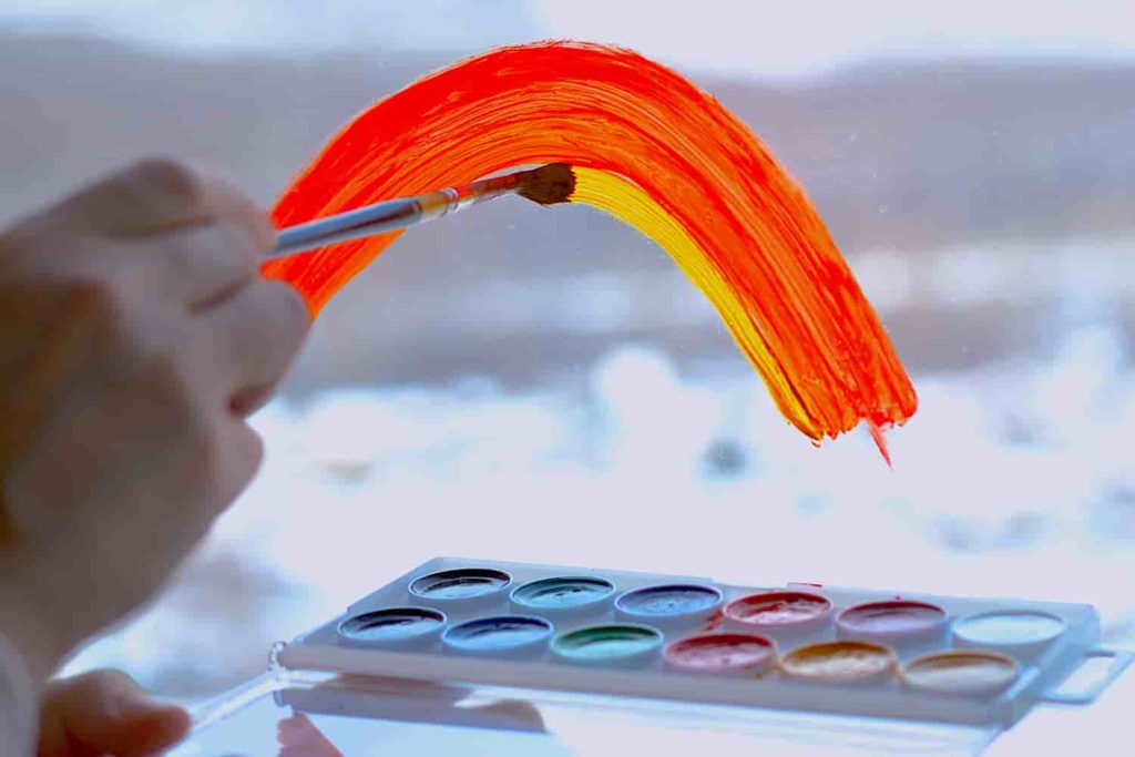A hand painting a rainbow on a window looking out on a snowy exterior.