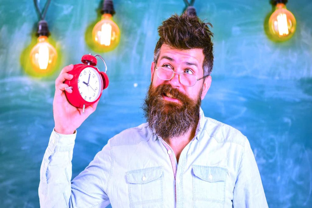 A bearded man with a distressed look holding an alarm clock in front of a chalkboard.