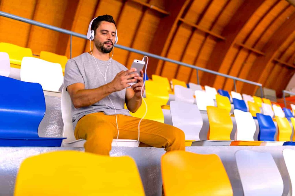 A man wearing headphones sitting by himself in bleachers looking at his phone.