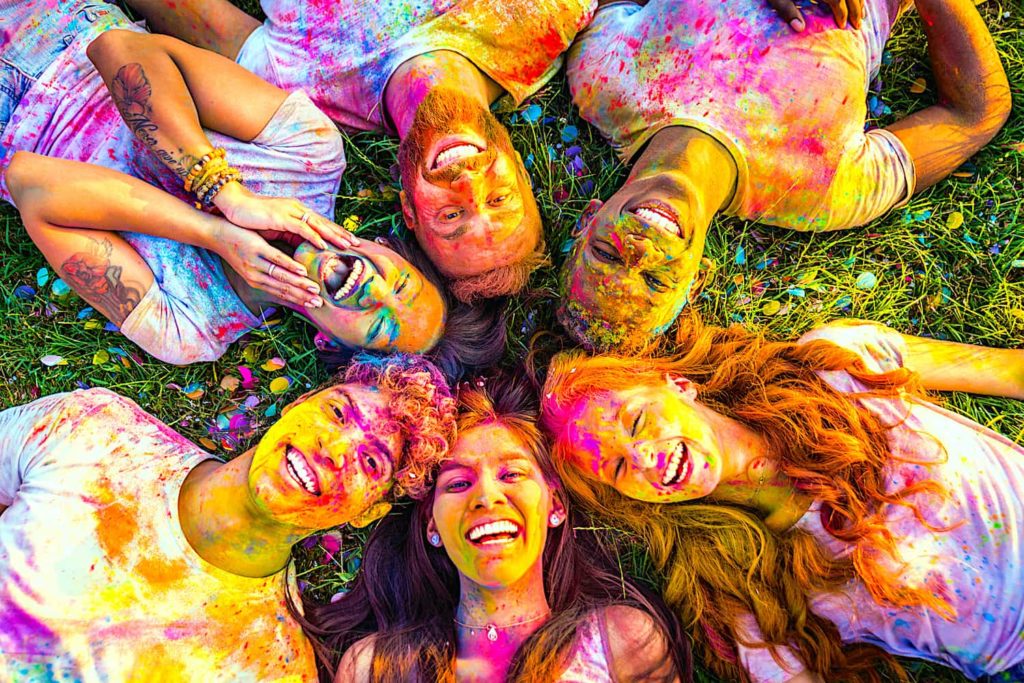Six people lying on the grass covered in colorful paint powder.