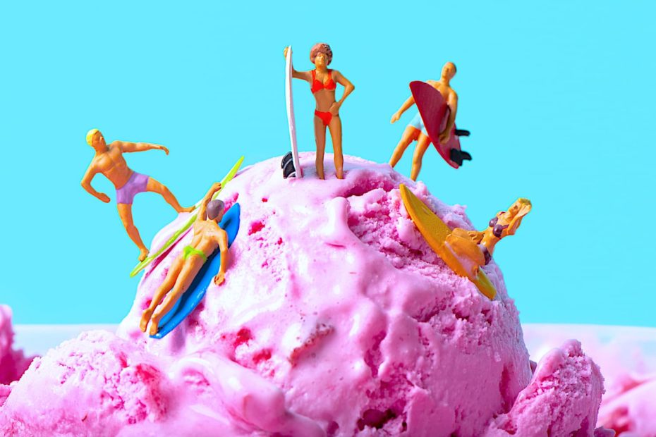 A scoop of pink ice cream with tiny surfer figures perched on the slopes of scoop.
